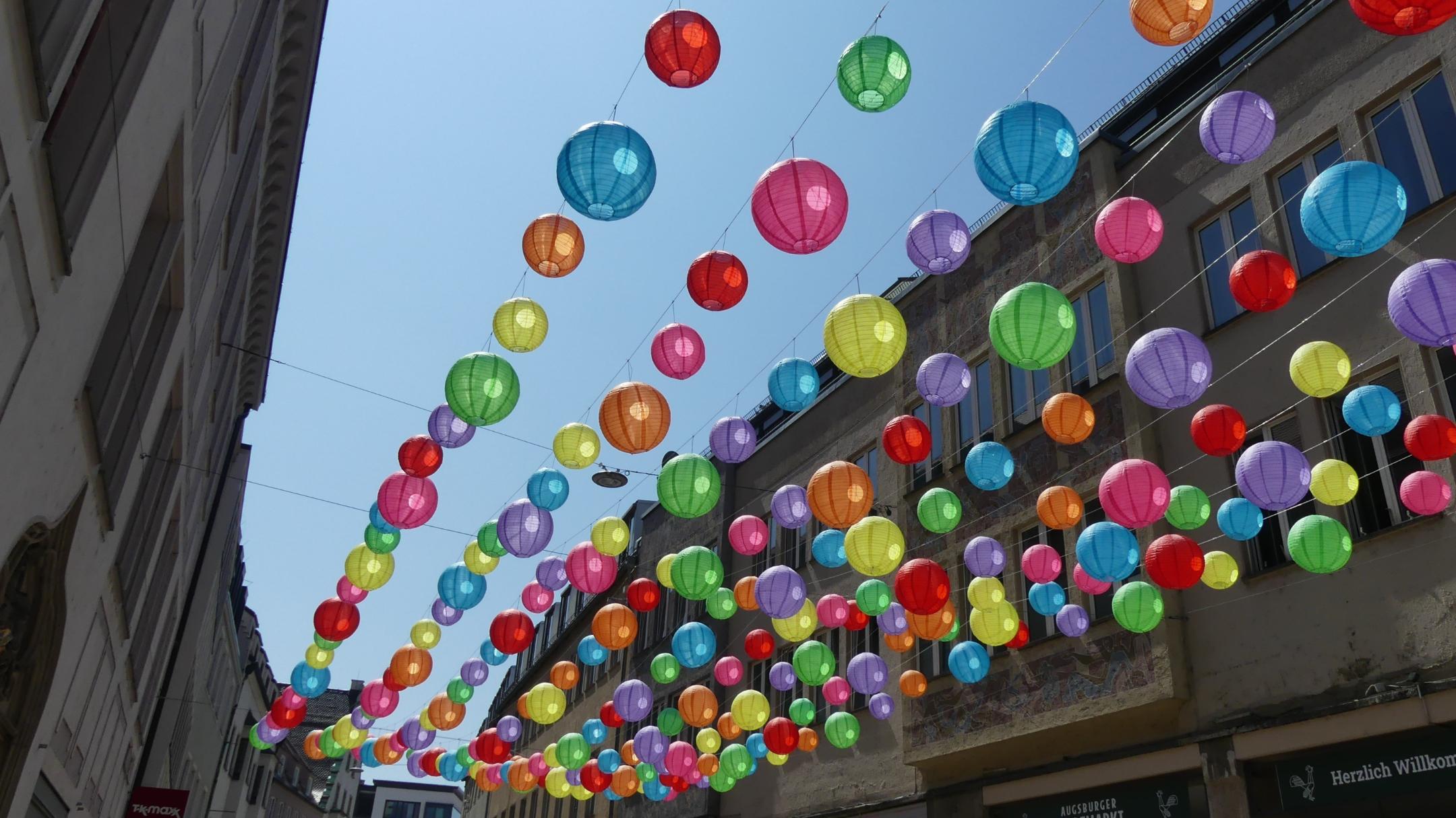 Bunte Lampions an Seilen aufgereiht in einer engen Straße im Hintergrund blauer Himmel